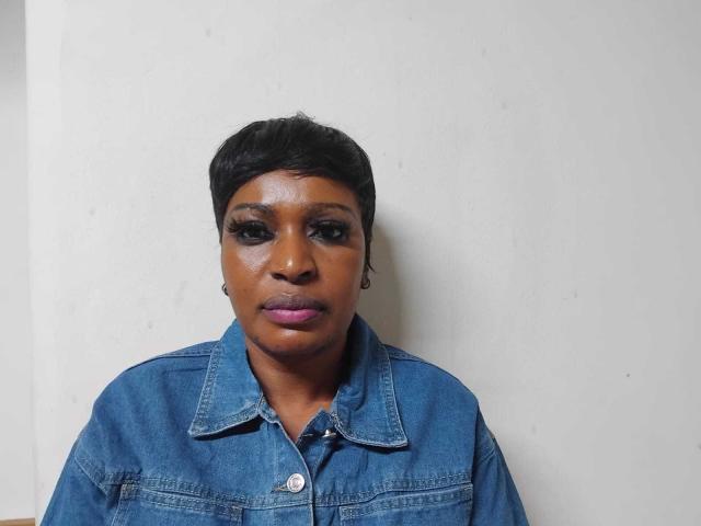 HotXBums with dark skin, short black hair, wearing a blue denim shirt, purple lipstick, and heavy eyelashes, standing against a white wall. Boobs not visible.