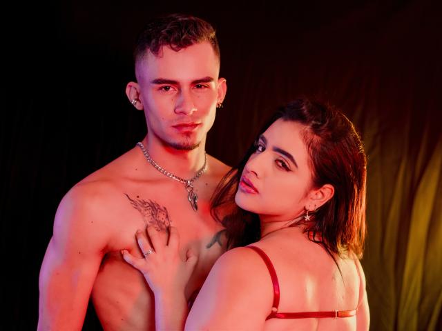 SharonAndTyga, a muscular shirtless guy with a tattoo and necklace, holds the hand of a red-brasied woman with dark hair, her hand on his chest. Red and pink lighting.