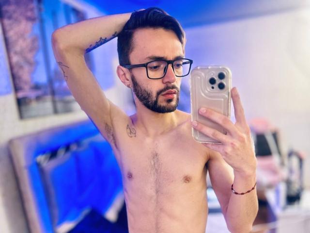 Shirtless JakeMagnus with glasses, beard, and tattoos, holding a phone, taking a mirror selfie, arm raised, showing a slim, hairy chest and a red bracelet.