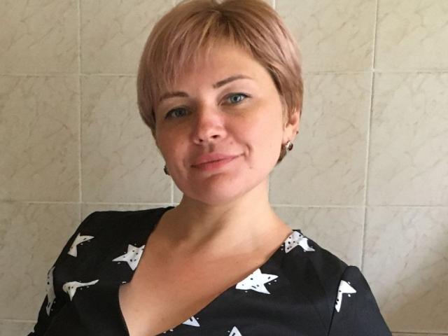 LadyElina has short pink hair, blue eyes, and a slight smile. She's wearing a black shirt with white star patterns, showing her tits. She's in a tiled room.