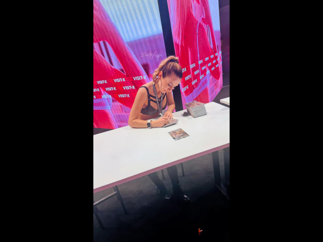 Lady-Anja with ponytail, black top, and necklace, leans over white table, looking at cards, wearing black boots, in bright pink and blue background.