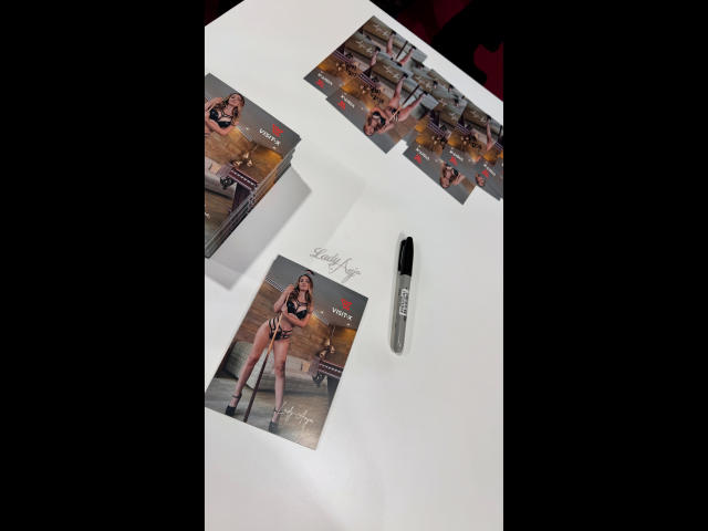Lady-Anja's tits on multiple glossy photos, black pen with "Wag" logo, white surface, top-down view, her legs spread, wearing black lingerie, posing in a wooden room.