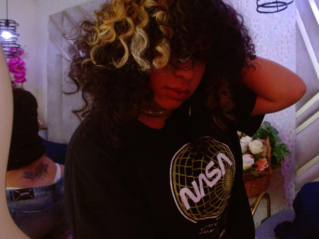 NeffellyRoss with curly black and blonde hair, wearing a black NASA t-shirt, hand in hair, choker, dark skin, nose ring, looking at camera, mirror reflection, pink flowers.