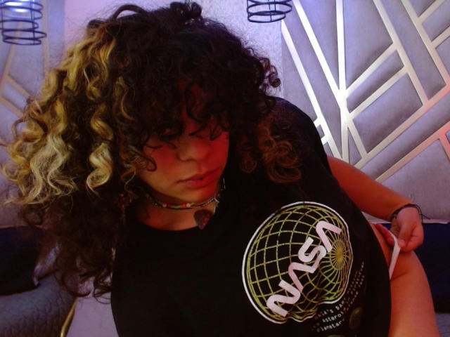 NeffellyRoss with curly black and blonde hair, wearing a NASA shirt, has her tits pushed up by her hand, looking down with a slightly pouty expression.
