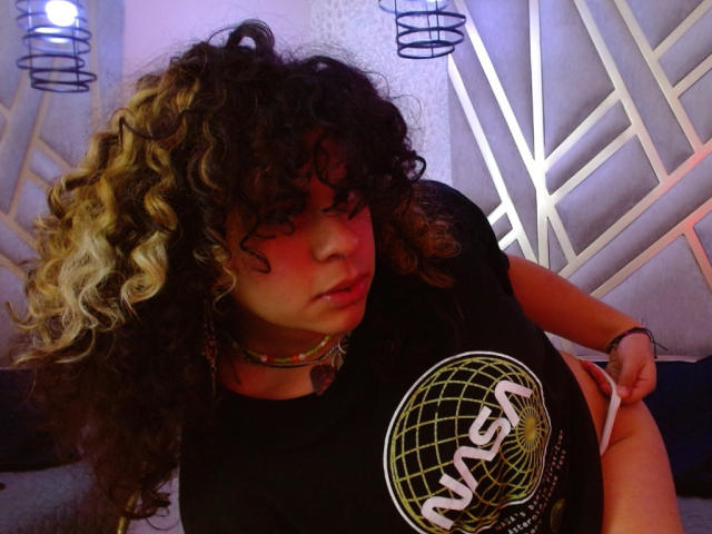 NeffellyRoss with curly black and blonde hair, wearing a black NASA shirt, pulling down white panties, exposing her ass, in a dimly lit room with blue and purple lights.