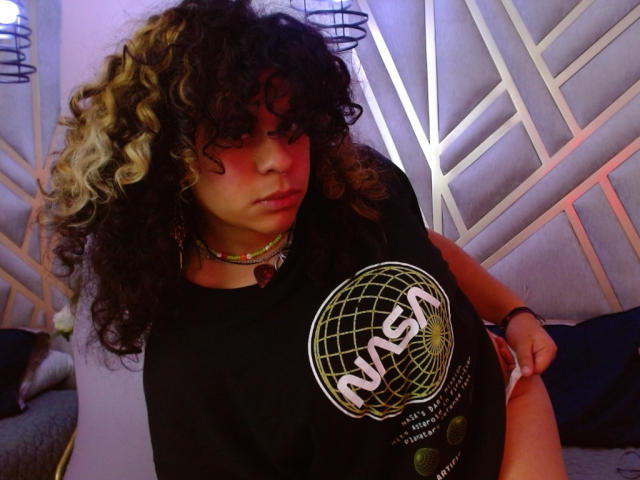 NeffellyRoss with curly black and blonde hair, wearing a NASA shirt, red choker, and colorful necklace, standing with a serious expression, slightly pulling down black pants.