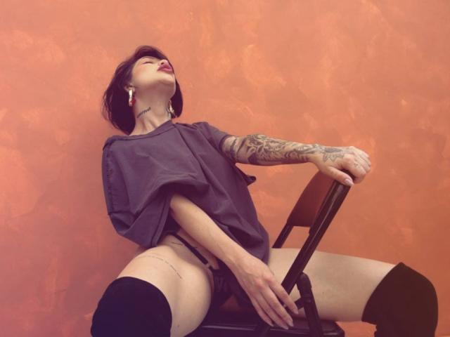 HannaBorbon, with tattoos on her arm, leans back on a chair, wearing a loose purple shirt, black thigh-high socks, and no pants, exposing her pussy. Red headphones in ear.