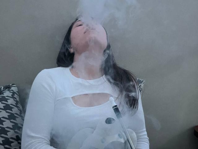 BelleEliza with eyes closed, inhaling thick smoke from a vaporizer, wearing a white cut-out top, her brown hair falling to the side, holding the vaporizer in her lap.