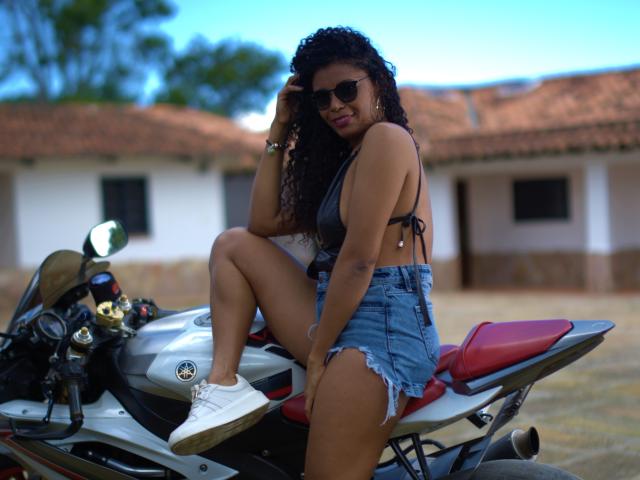 AnelicaSexos with curly black hair, wearing a black bra and high-waisted denim shorts, sits on a silver motorcycle with red seats, showing her white sneakers and brown skin.