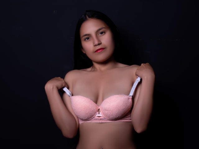 MelanieGates, with dark hair and red lipstick, holds pink lace bra straps, showing her tits against a black background. She has a small nose ring and a confident look.
