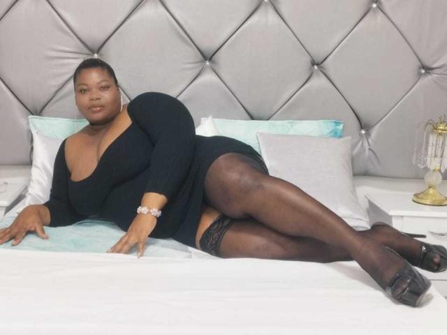 MilenaLopez, curvy black woman, laying on bed, wearing black dress, black stockings, black heels, bracelet, butt raised, legs crossed, looking at camera, tufted headboard, pillow, white bed.