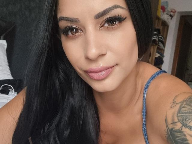 SweetyFinger has long black hair, brown eyes, and glossy pink lips. She's wearing a blue spaghetti strap top, showing a tattoo on her right shoulder. Her face is shiny.
