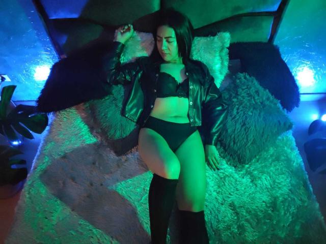 ShairaBrunette está acostada en una cama, vistiendo una chaqueta de cuero negro, un sujetador negro y ropa interior negra, con luces verdes y azules, y calcetines hasta la rodilla negros, con las piernas ligeramente separadas..