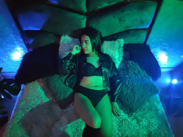 Shaira, una morena con un brillo verdoso, acostada en una cama, vistiendo un sujetador y tanga negros, una chaqueta de cuero negro, calcetines largos oscuros y almohadas suaves, mirando hacia un lado..