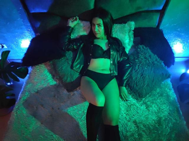 Shaira: Una morena con cabello oscuro, chaqueta de cuero negra, sujetador negro, ropa interior negra y medias negras hasta la rodilla, acostada sobre almohadones suaves, con luces verdes y azules, una pierna levantada, mirando a la cámara..