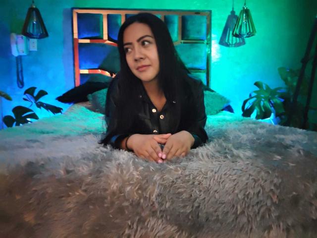 Shaira, una morena con cabello negro, vestida con una camisa negra, con las manos sobre una manta gris suave, mirando ligeramente hacia un lado con una sonrisa sutil, en un dormitorio iluminado con luz azul..