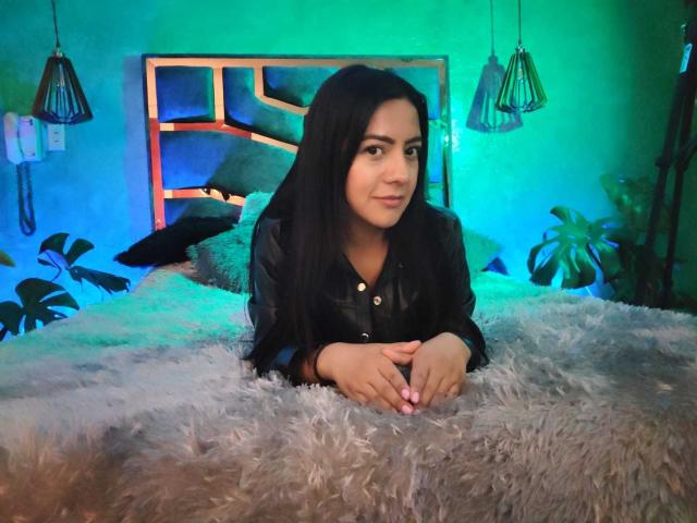 Shaira, una morena con cabello largo y negro, una camisa de cuero negro y uñas rosas, acostada en una cama gris y esponjosa, con las manos juntas, y luces verdes y azules en el fondo..