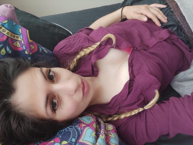 SkylerSamuel lying on a couch, wearing a purple shirt, bra visible, blonde braids, fair skin, brown eyes, small tits, relaxed, hand on belly, colorful pillow, casual.