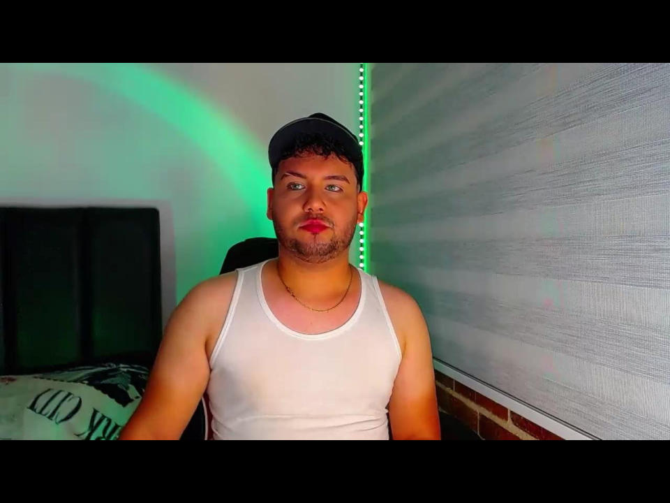 SexyMenX69, wearing a white tank top and black cap, with a beard and red lipstick, standing against a wall with green lighting in the background.