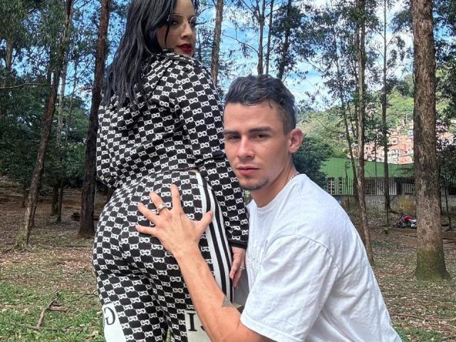 SharonAndTyga with black and white patterned pants, butt in face, standing, holding it. Tyga, white t-shirt, short brown hair, looking up. Forest background, blue sky.