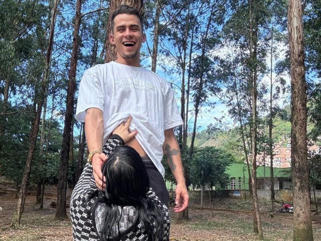 SharonAndTyga with a white t-shirt, smiling, standing in a forest. A woman with black hair, wearing a black and white patterned top, is lifting his shirt, showing his abs.