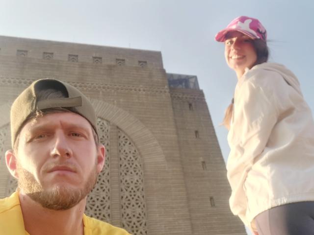 Two people, sunlit, in front of a brick building. MissmeanDre with brown beard, green cap, yellow shirt. Another MissmeanDre, pink hat, white jacket, smiling.