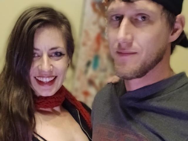 MissmeanDre with tits showing in a black top, red scarf, and smiling, next to a guy in a grey shirt, black cap, and slight beard, both smiling.
