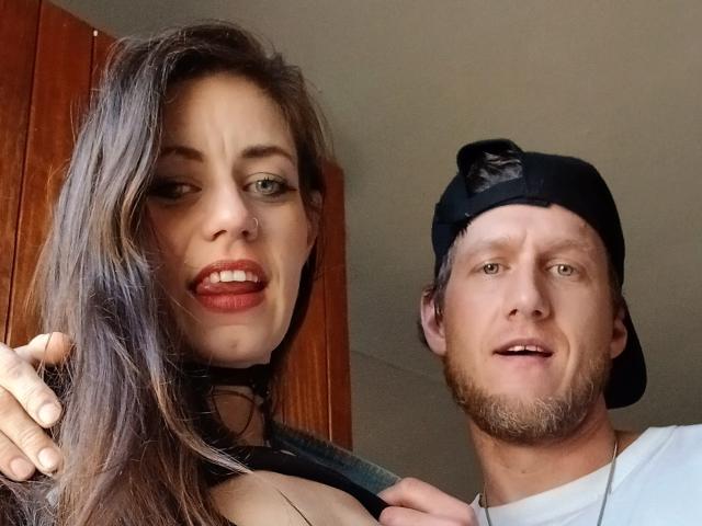MissmeanDre with long brown hair and red lipstick, showing her tits, next to a bearded guy in a black cap, smiling. She's on the left, he's on the right.