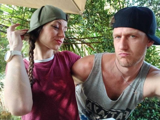 MissmeanDre with braided brown hair, green cap, red shirt, and watch, next to a serious-looking man with a black cap, grey tank top, and necklace.