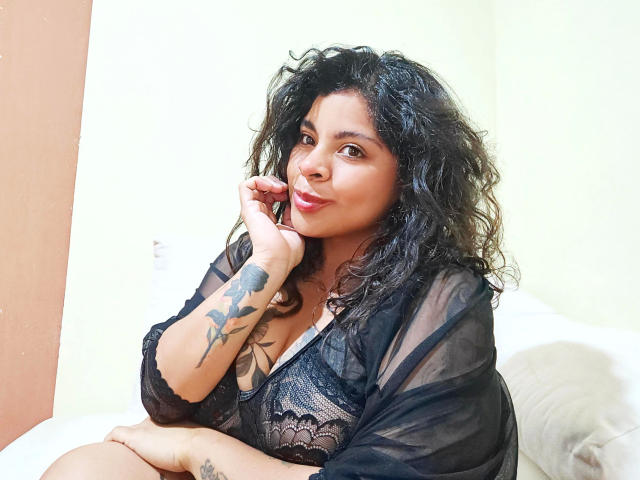 SexyPraline-hot with curly black hair, dark skin, wearing black lace lingerie and sheer robe, smiling, resting chin on hand, tattoo of bird on left arm, sitting on bed.