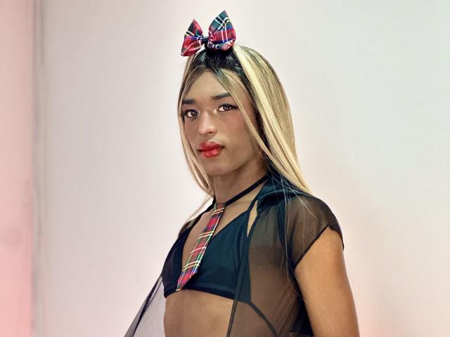 ParisGlamour with blonde hair, red lips, and a plaid bow, wearing a sheer black top, exposing small tits, and a colorful tie. She has a cute, confident expression.