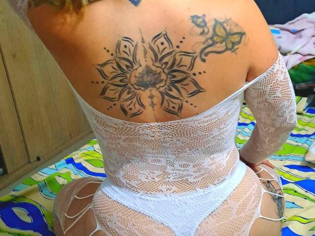 PerlaGoloza has a large, intricate tattoo on her back, wearing a white lace bodysuit that exposes her ass. Her blonde hair is visible, tattoos are on her upper back.