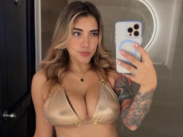 AnastasiaDolly with blonde wavy hair, wearing a shiny gold bikini top, showing her big tits and tattooed arm, taking a selfie with a white phone in a bathroom.