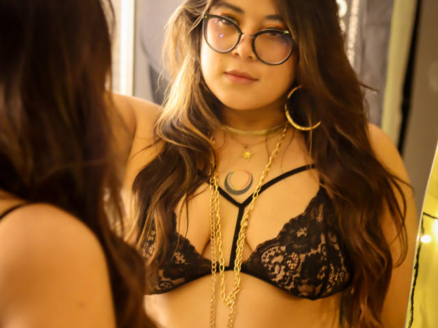 IsabelArt with wavy brown hair, glasses, and gold hoop earrings, wearing a black lace bra, looking at herself in a mirror. Gold necklace and crescent moon pendant visible.