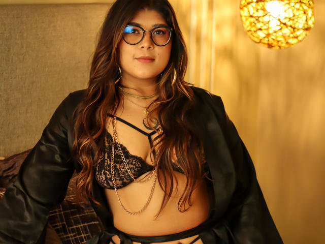 IsabelArt with long brown hair, glasses, black lace bra, chain necklace, and open black robe, showing her tits, sitting in a warm, yellow-lit room.