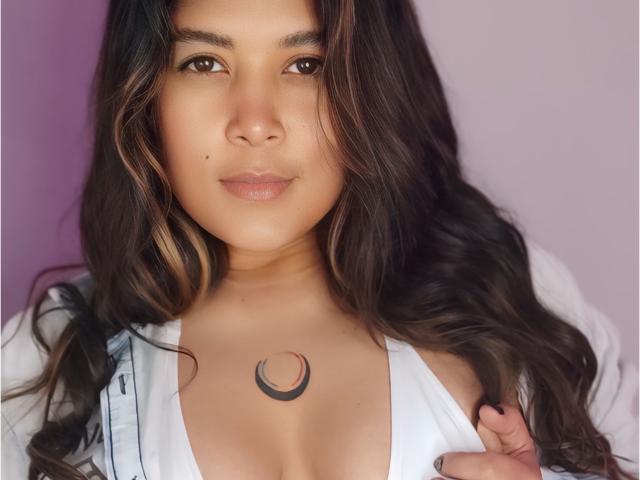IsabelArt with long brown hair, tan skin, and a crescent moon tattoo on her chest, wearing a white shirt, slightly pulling it down to show cleavage, with a neutral expression.