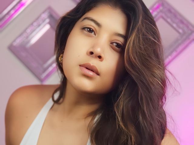 IsabelArt with wavy brown hair, light brown skin, and pouty lips in a white top, looking slightly to the side with a relaxed, seductive expression. Boobs partially visible.