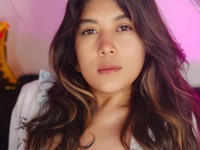 IsabelArt with wavy brown hair, brown eyes, and a small beauty mark on her right cheek, wearing a white top, showing a subtle tattoo on her chest.