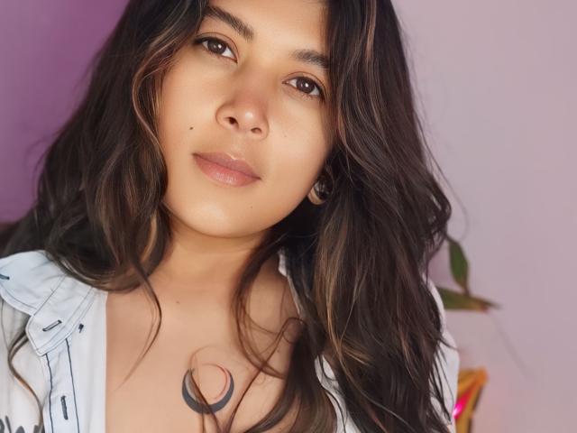IsabelArt has long, wavy brown hair, a cute nose, and a small tattoo of a crescent moon on her chest. She's wearing a white, unbuttoned shirt, looking at the camera.