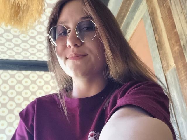 BelleEliza with glasses, brown hair, and a maroon shirt, smiling slightly at the camera, showing a small bit of her arm. Her tits are small, and she's cute.