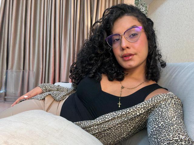 Rachell Beaumont mit lockigem, schwarzem Haar, die eine Brille trägt, ein schwarzes Oberteil, einen Leopardenmuster-Cardigan, eine Kreuzkette und beige Hosen, sitzt auf einem grauen Sofa mit einem weißen Kissen..