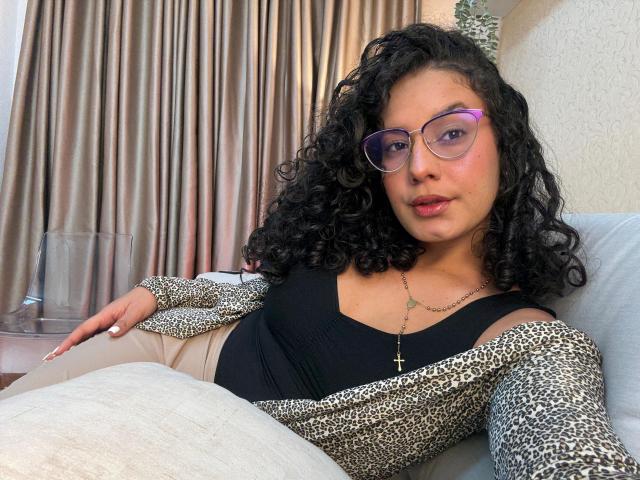 RachellBeaumont with curly black hair, purple glasses, black tank top, leopard cardigan, cross necklace, beige pants, lying on couch, looking at camera, soft smile, pink lips.