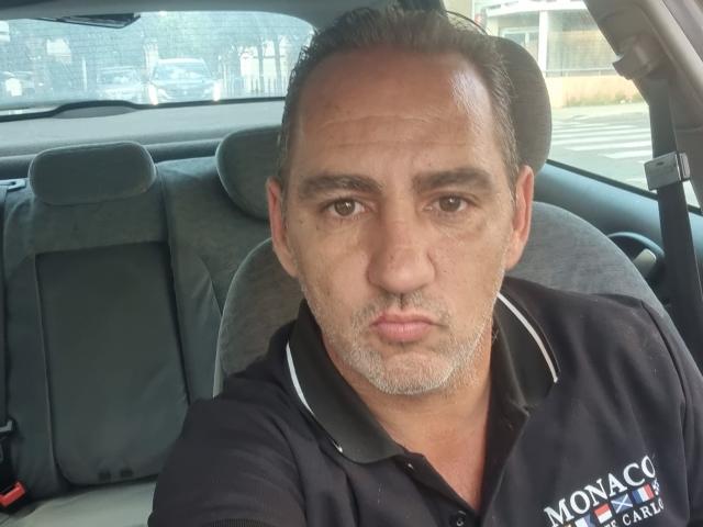 ManiSexy69 has a serious face, short dark hair, a light brown beard, and is wearing a black polo shirt, sitting in a car. His brown eyes look straight at the camera.