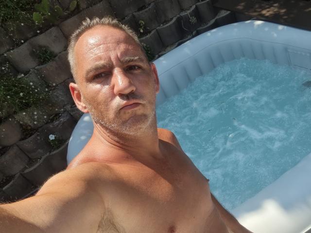 ManiSexy69, middle-aged with shaved head and light beard, shirtless, standing in a blue inflatable hot tub, looking up at the camera with a serious expression.