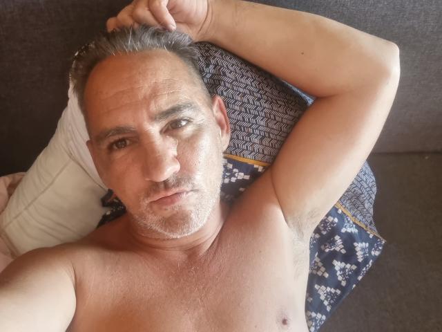 Shirtless ManiSexy69 with gray hair, lying on a pillow, arm raised, showing armpit, looking at camera, slightly wrinkled skin, casual expression, sitting on gray couch.