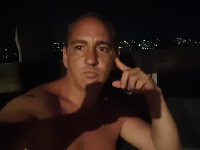 Shirtless ManiSexy69 with a serious expression, pointing at his right temple with his index finger. Bald with a slight tan, dark night background with city lights.