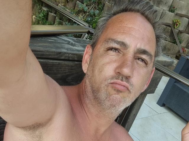 Shirtless ManiSexy69 with gray hair and stubble, holding up phone for selfie, showing armpit hair and slightly puffy ass, looking into camera with a neutral expression.