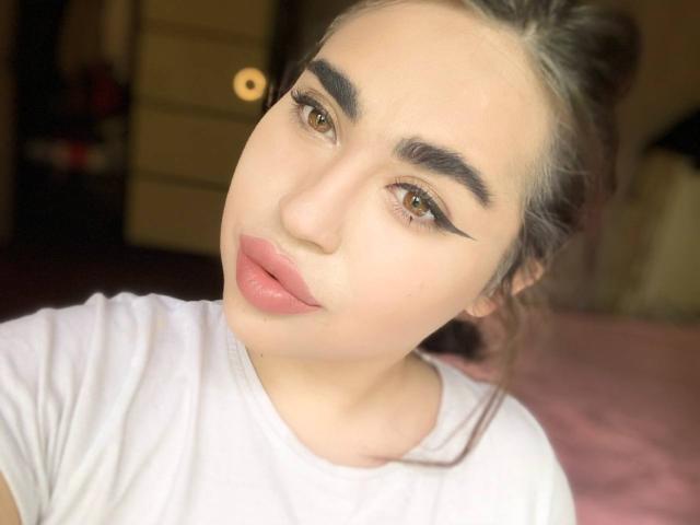 MeganWoww with pouty pink lips, thick black eyebrows, and winged eyeliner, wearing a white shirt, has brown eyes and light skin, with gray hair in a messy bun.