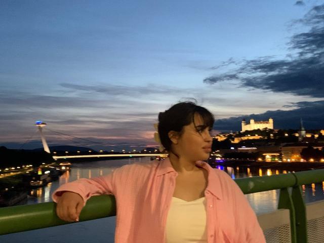 MeganWoww with dark hair in a pink shirt and white top, leaning on a green railing at dusk, a tower and illuminated building in the background.