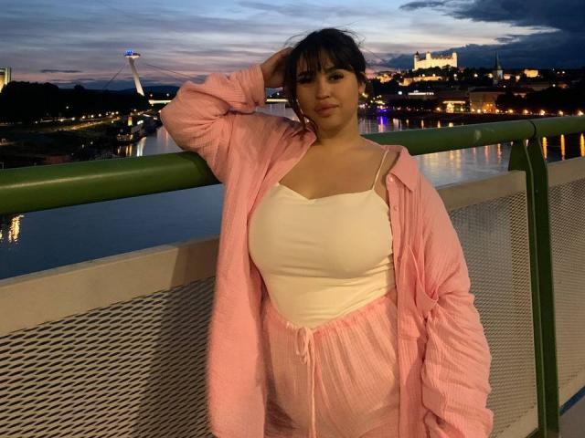 MeganWoww with her tits showing in a white tank top, pink pajama pants, and pink jacket, leaning on a green railing at dusk, hand in her hair.